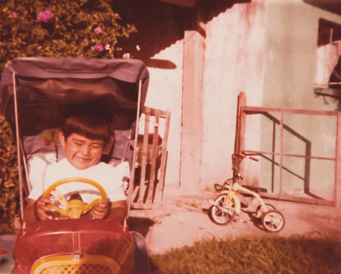 Fotografía antigua de Esmeralda Ahuatzi, CEO y fundadora de Kreaz DG, sonriendo mientras juega en un auto de juguete rojo con toldo, junto a un triciclo amarillo en el patio de una casa.